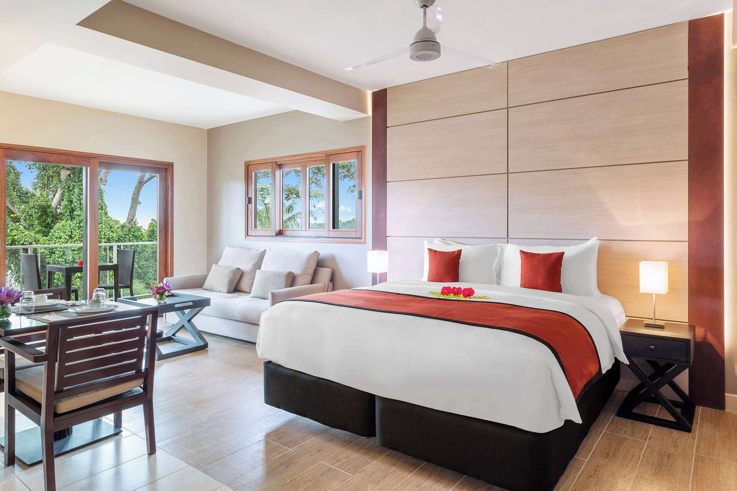 A modern hotel room with a large bed, a sofa, a dining table, and a balcony with a view of greenery.