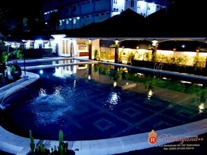 A swimming pool with a curved edge, surrounded by a landscaped area with plants. There are buildings in the background, and a sign with the hotel's name and contact information is visible. - Ramayana Tasik (Tasikmalaya)