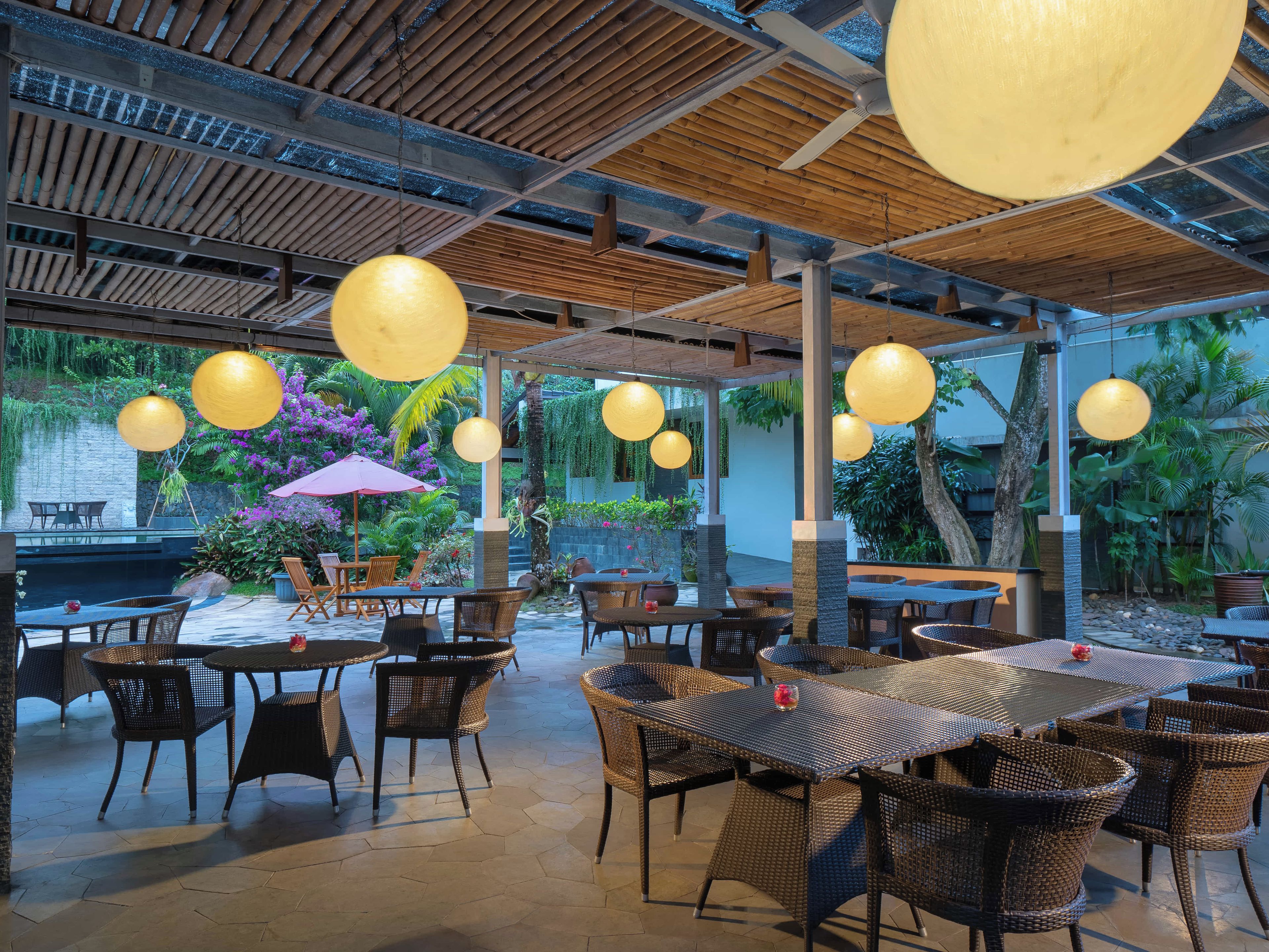 An outdoor dining area with wicker furniture, hanging spherical lights, and a view of a garden with tropical plants.