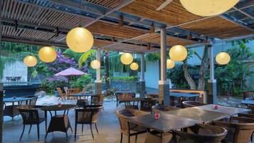 An outdoor dining area with wicker furniture, hanging spherical lights, and a view of a garden with tropical plants.