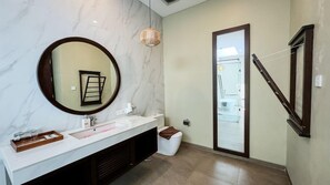 A modern bathroom with a marble vanity, a round mirror, a hanging light fixture, and a glass shower enclosure. - Rumah Kito Jambi (Jambi)