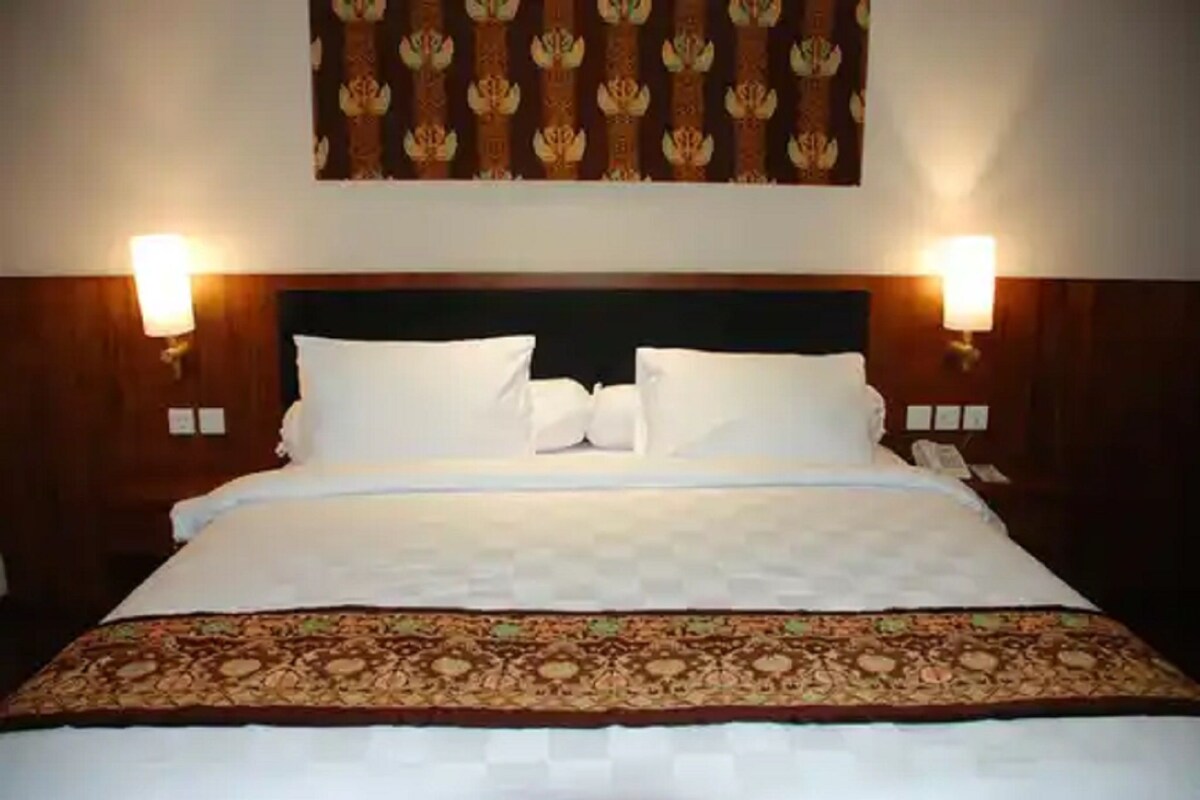 A hotel room with a large bed, two bedside lamps, a patterned headboard, and a floral wall hanging.