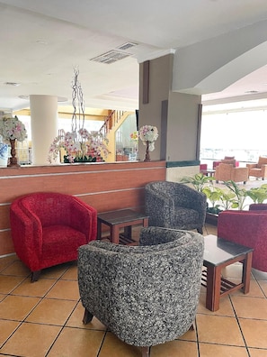 Eine Lobby mit einer Rezeption, roten und gemusterten Sesseln, einer Treppe und Blick ins Freie.