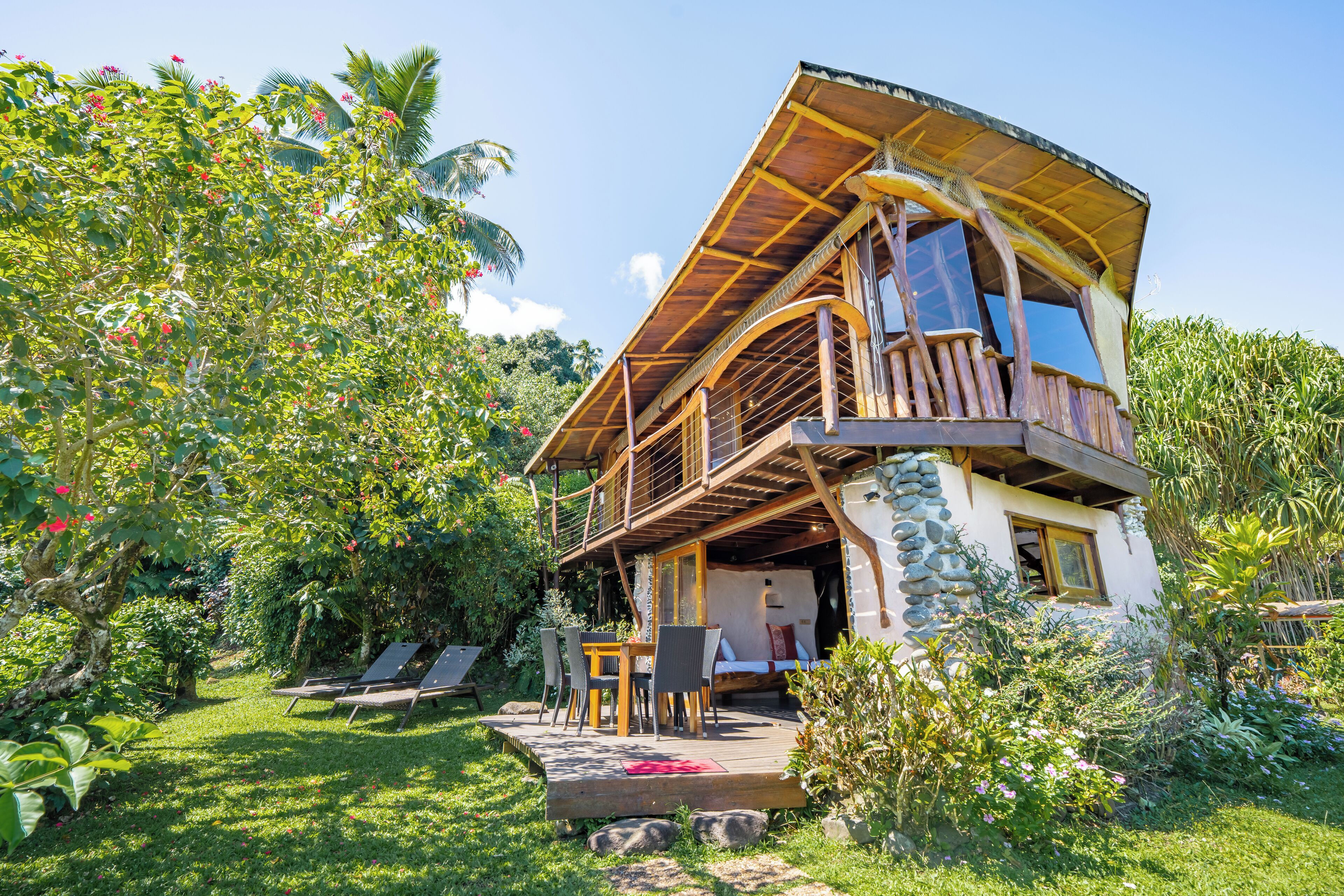 Una casa a due piani in legno con un balcone, circondata da vegetazione rigogliosa e un giardino.