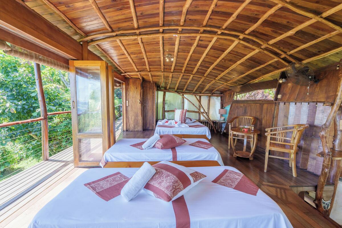 Une cabane en bois avec trois lits, chacun agrémenté de draps blancs et de touches de rouge, un plafond en bois et un balcon offrant une vue sur la verdure.