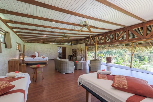A spacious room with wooden beams, a ceiling fan, a bed, a sofa, a small table, and a view of greenery outside. - Vanira Lodge (Taiarapu-Ouest)