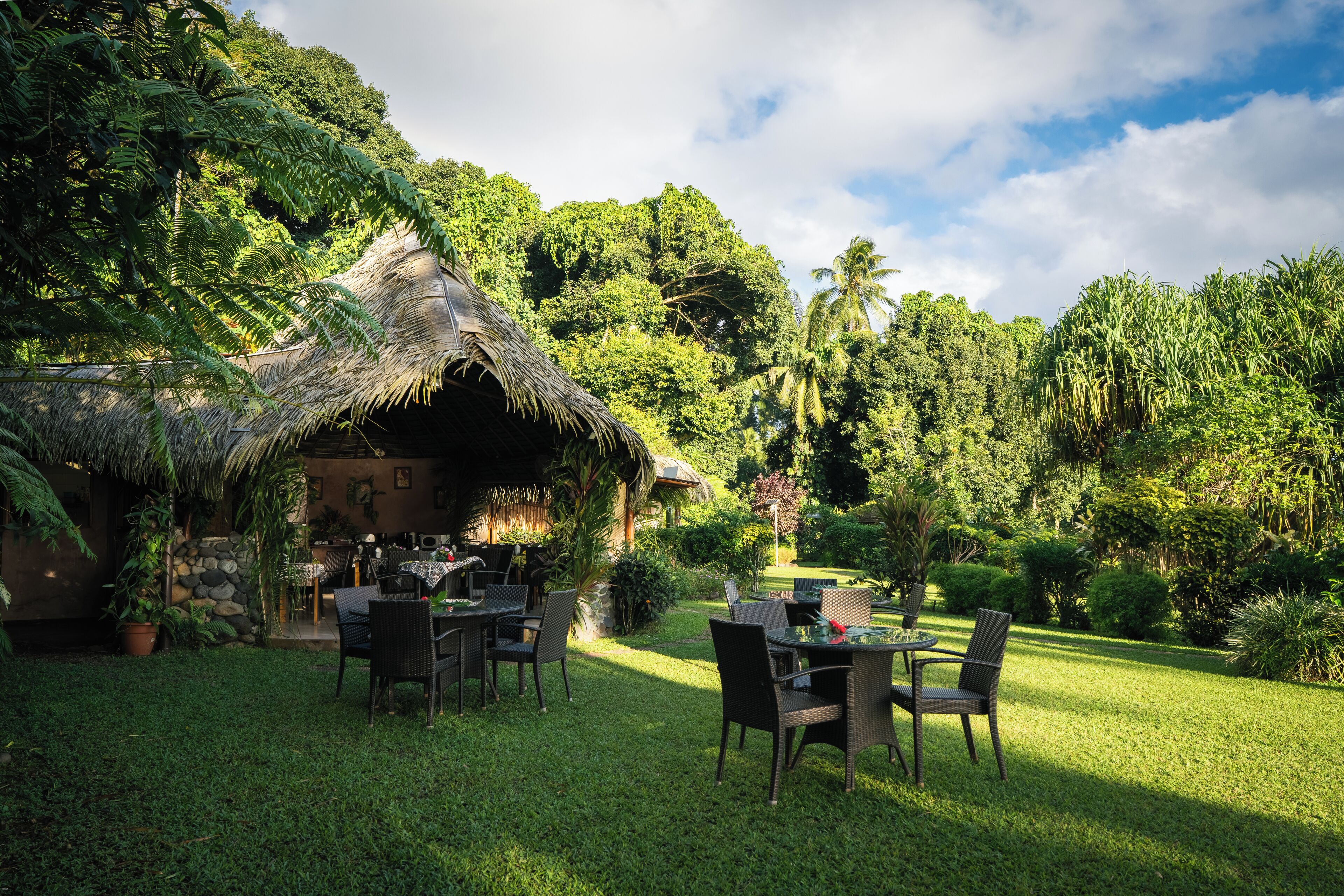 A thatched-roof outdoor dining area with wicker furniture, surrounded by lush greenery and a stone wall.