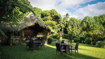 A thatched-roof outdoor dining area with wicker furniture, surrounded by lush greenery and a stone wall.