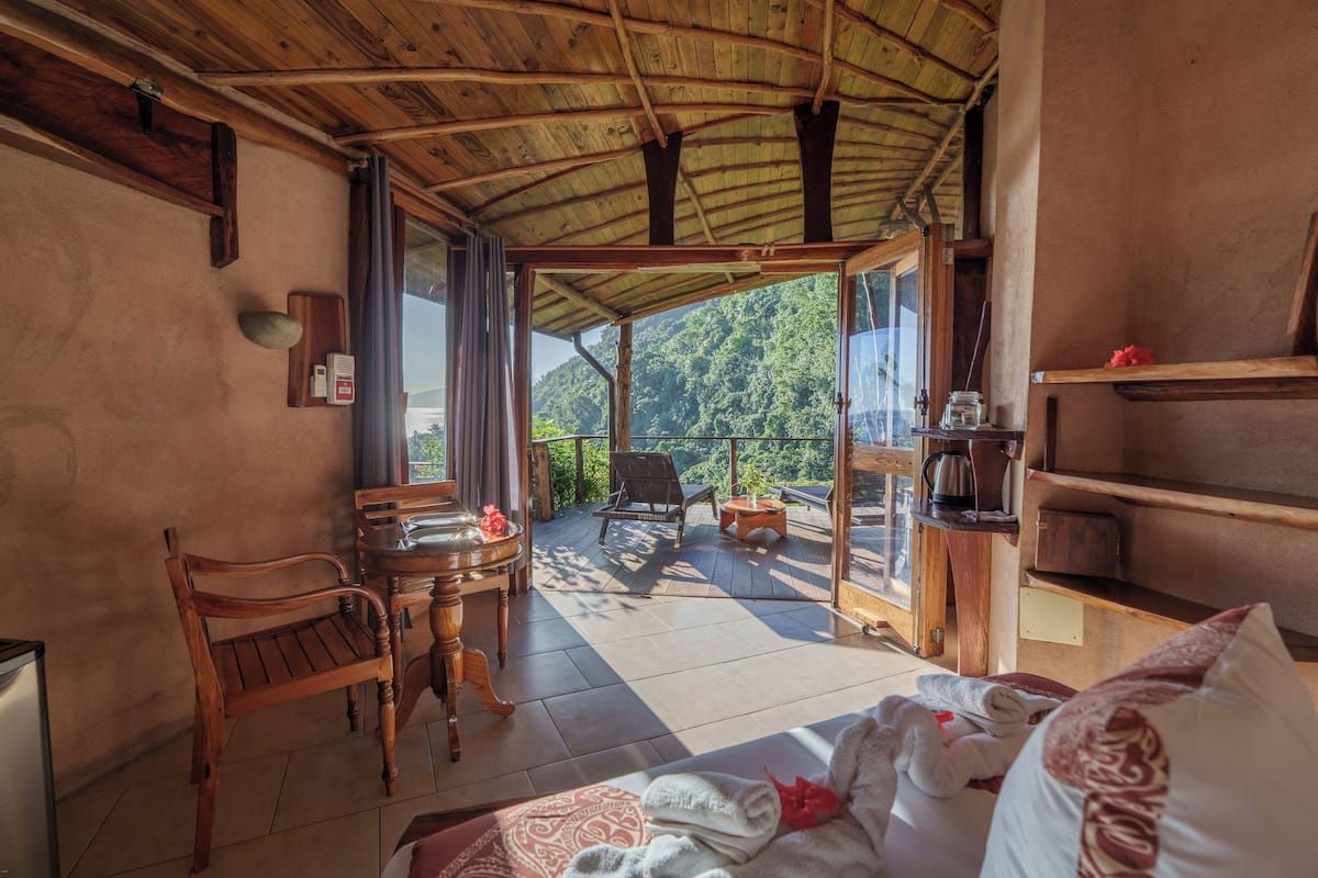 Une pièce avec un plafond en bois, un lit recouvert d’une couverture à motifs, une chaise en bois, une petite table avec une bouilloire, et une vue sur une zone boisée, visible à travers une porte vitrée.