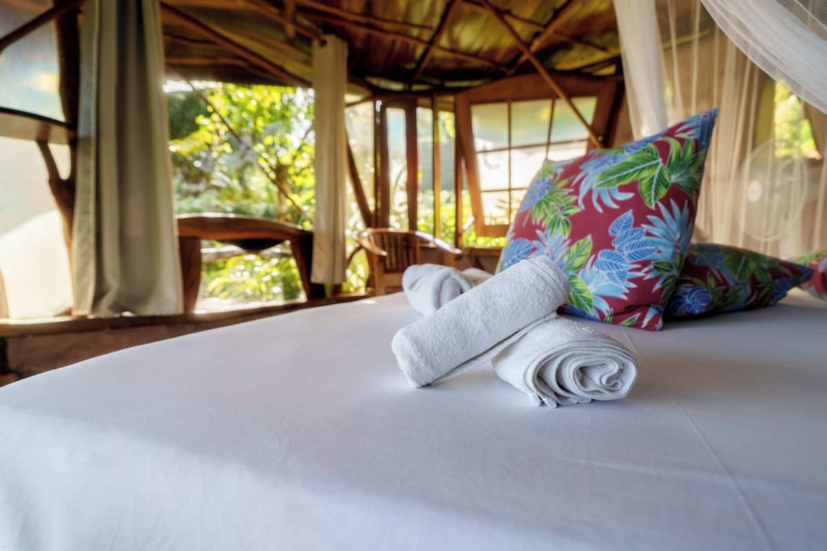 Un lit bien fait, avec une serviette blanche roulée et un coussin à motifs floraux, une chaise en bois et une vue sur la verdure par la fenêtre.