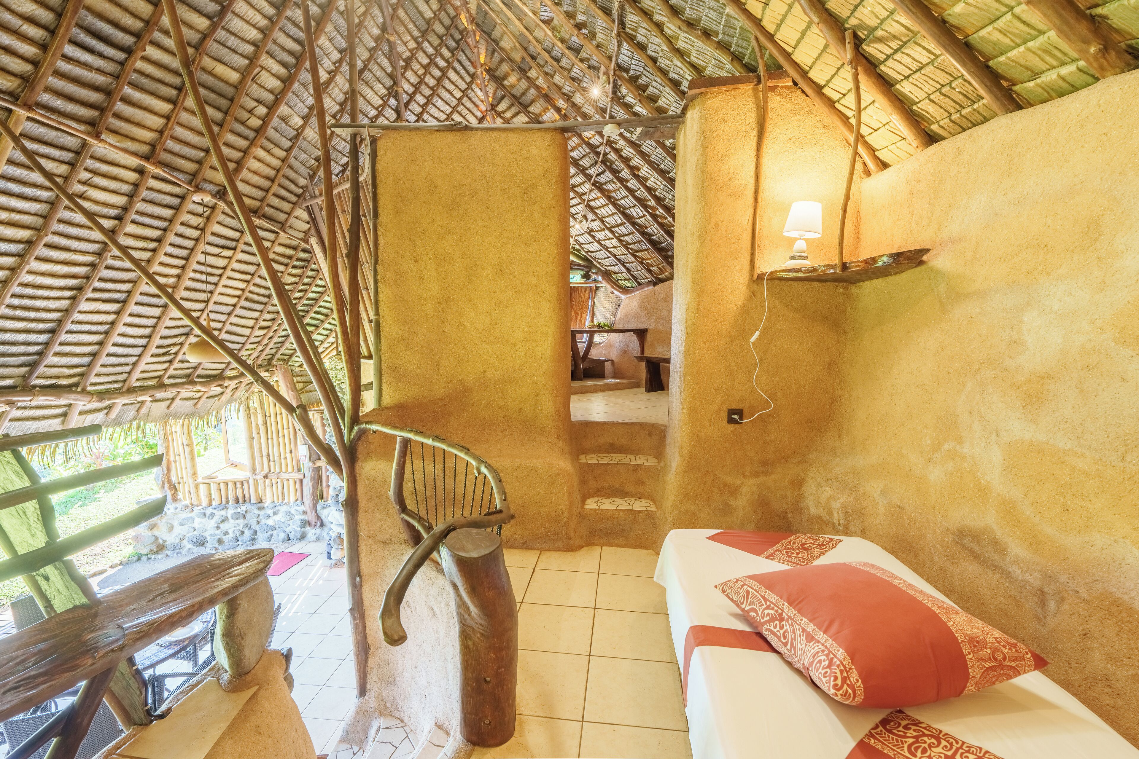 A rustic room with a thatched roof, a staircase, a bed with red and white bedding, and a small shelf with a lamp.