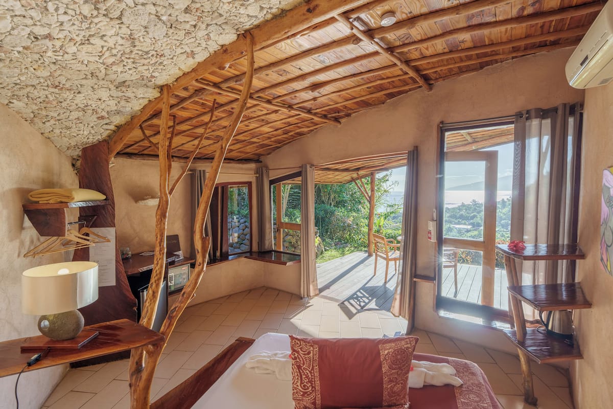 Une pièce avec un plafond en pente, des poutres en bois, un lit avec un oreiller rouge, un bureau avec une lampe et une vue sur la verdure à l’extérieur.
