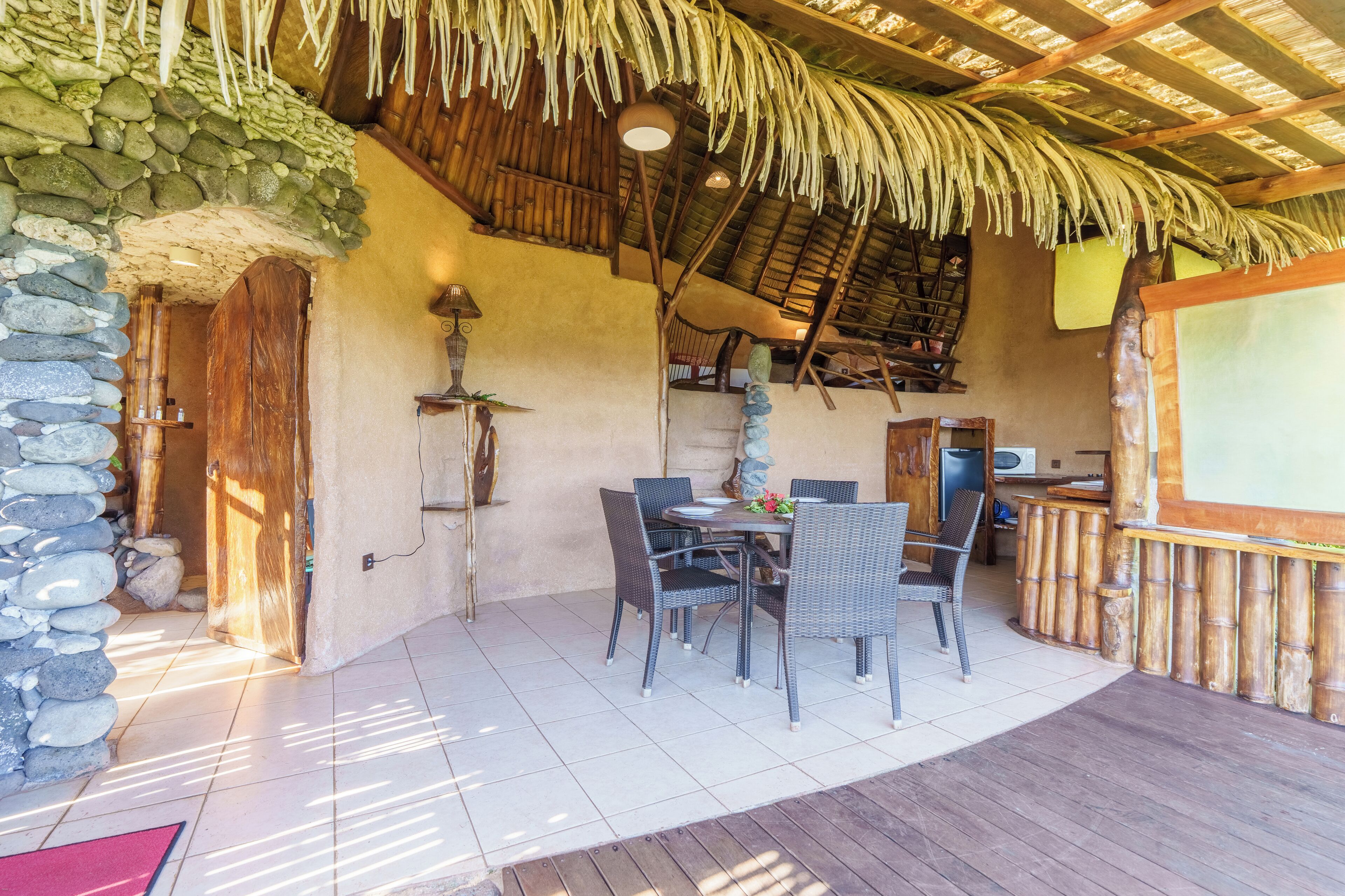 A rustic outdoor dining area with stone walls, a thatched roof, and a wooden deck.
