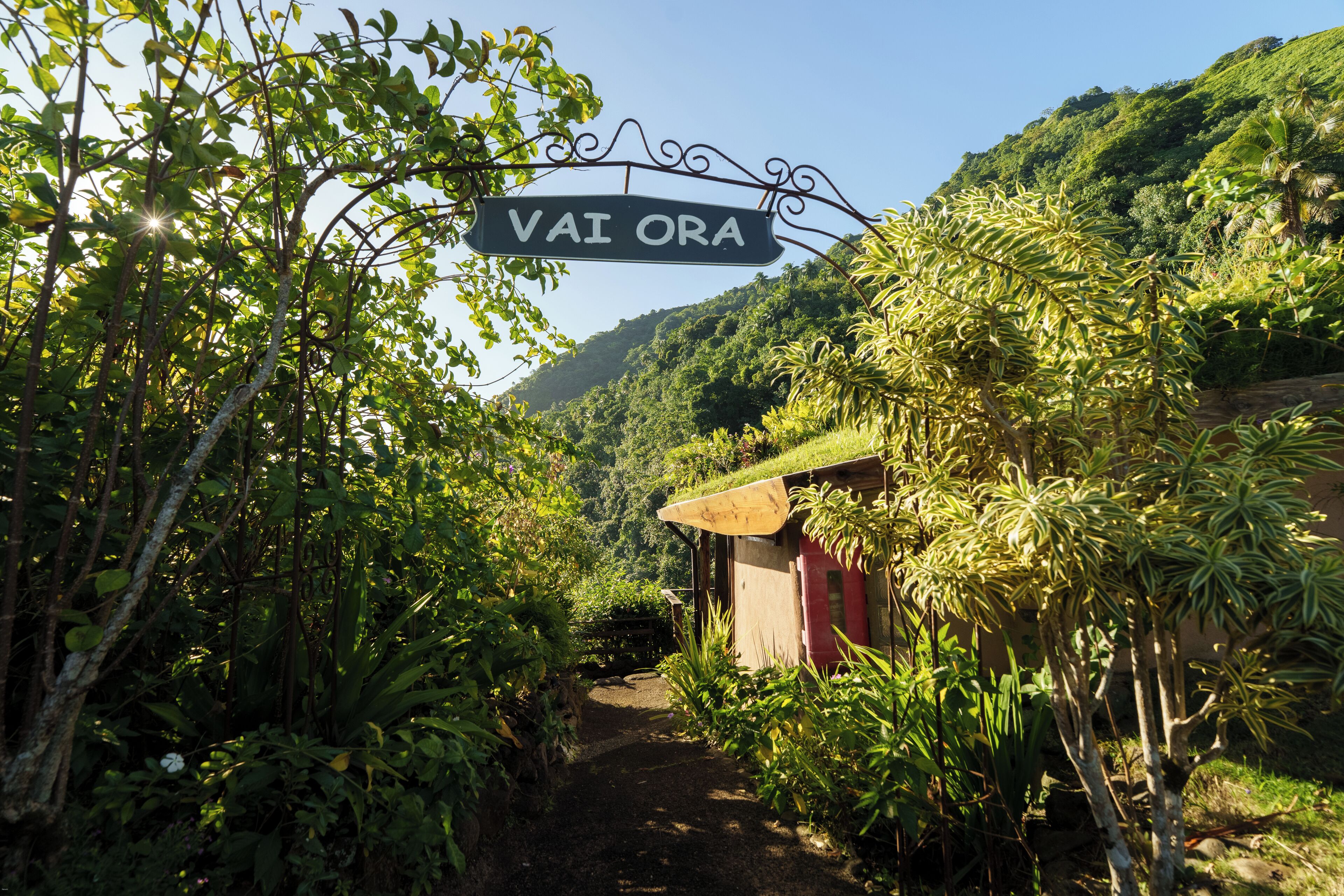 A sign reading "VAI ORA" in a lush, green environment with a path leading to a small building.