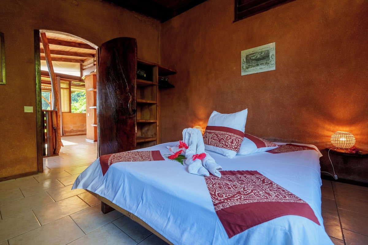 Un lit bien fait, recouvert d’une couvre-lit à motifs rouges et blancs, avec une tête de lit en bois et une table de chevet surmontée d’une lampe.