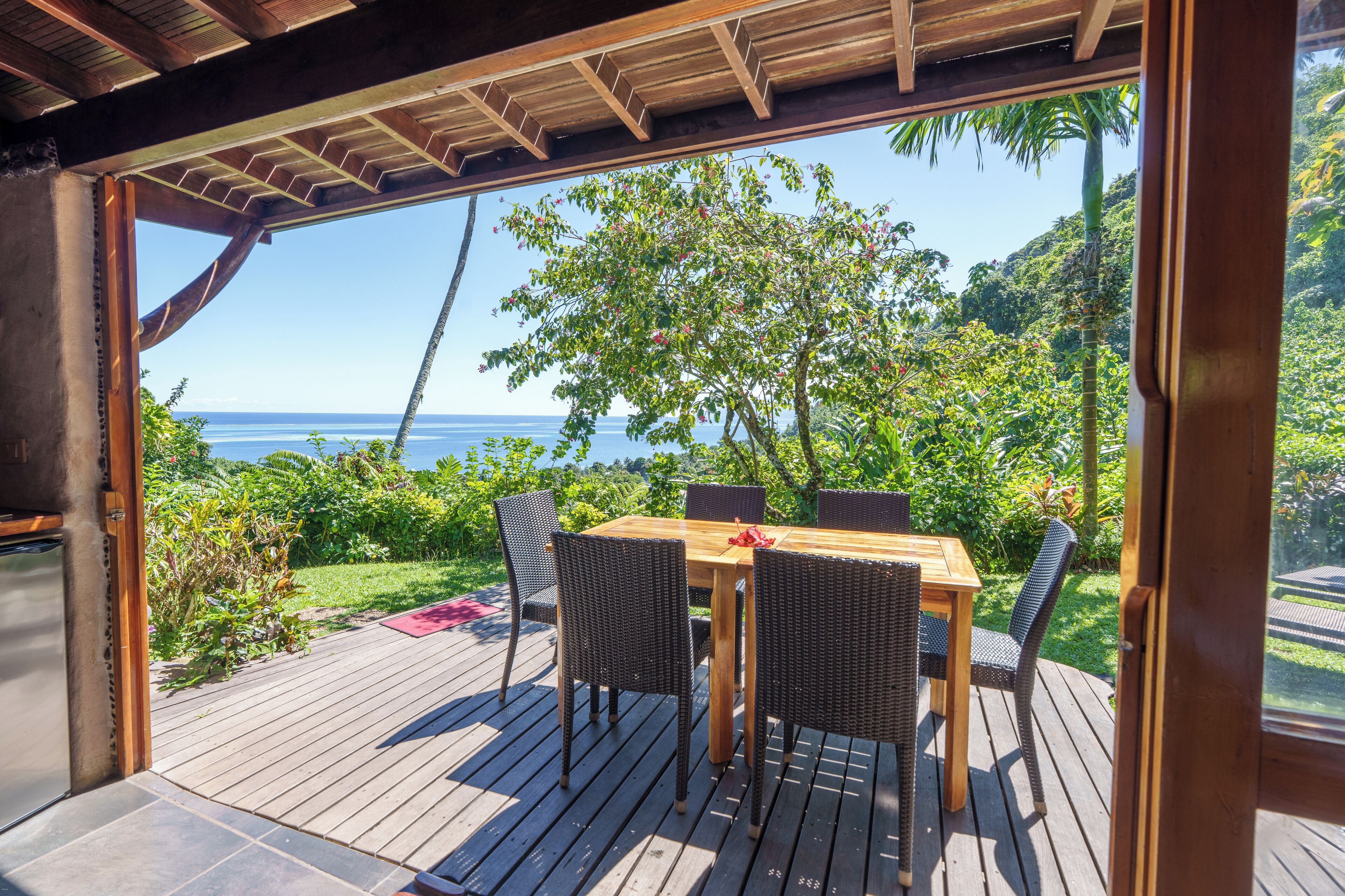 An outdoor wooden deck with a table and chairs, overlooking a lush garden and the ocean.