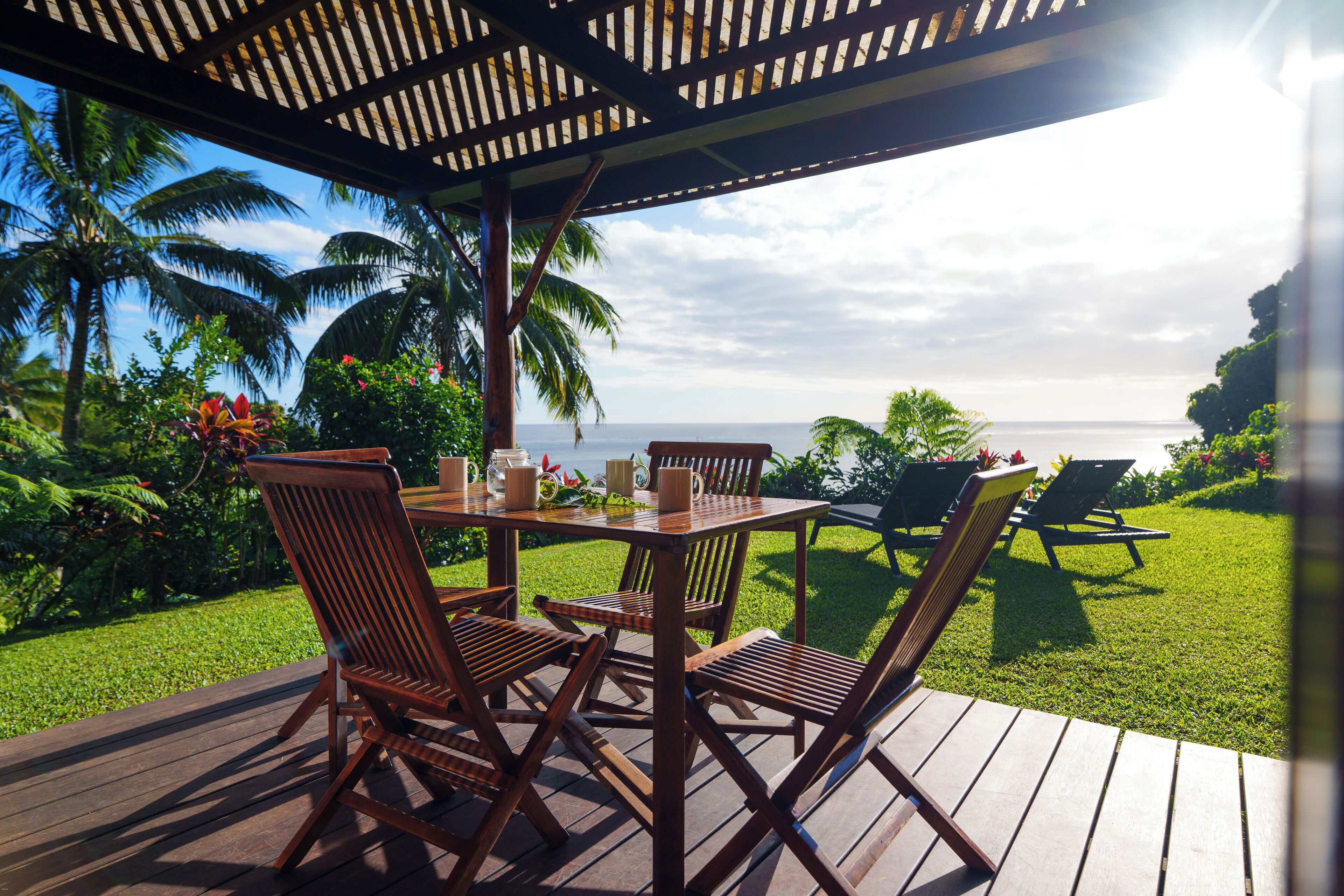 Um deck de madeira com mesa e cadeiras, com vista para um jardim exuberante e o oceano.