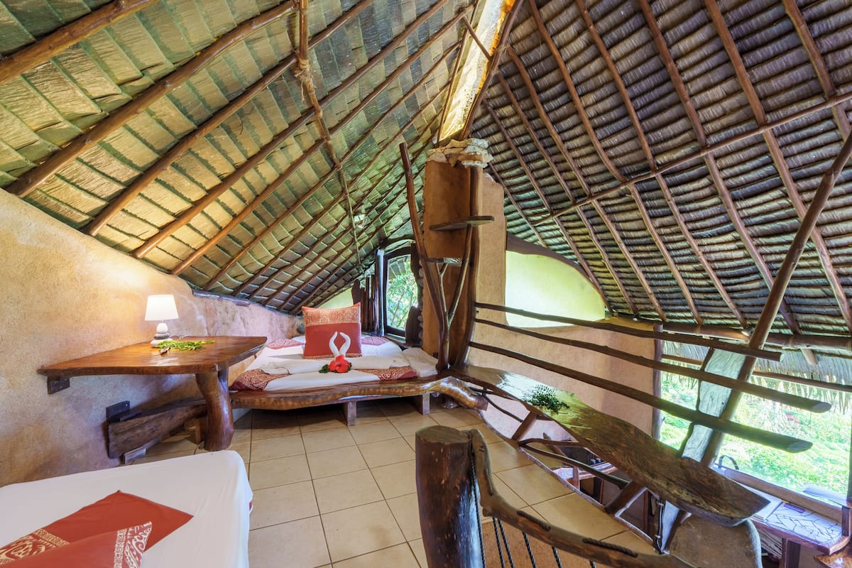 Une pièce avec un toit en pente, un lit avec un oreiller, une table avec une lampe et une fenêtre donnant sur la verdure.