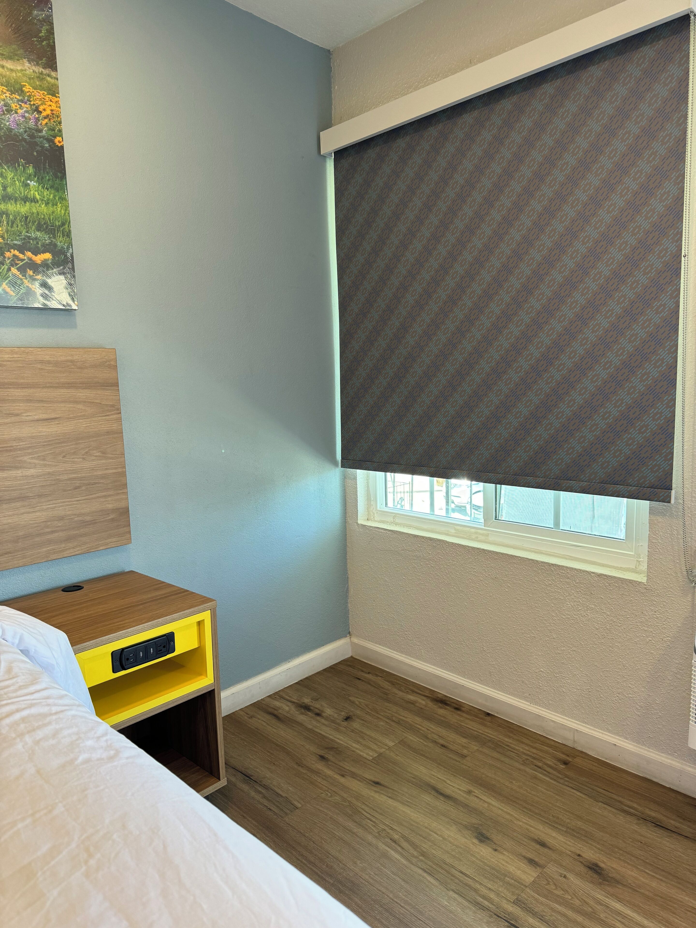 Un dormitorio con cama, una mesita de noche con toma de corriente, una ventana con persiana y un estante en la pared.
