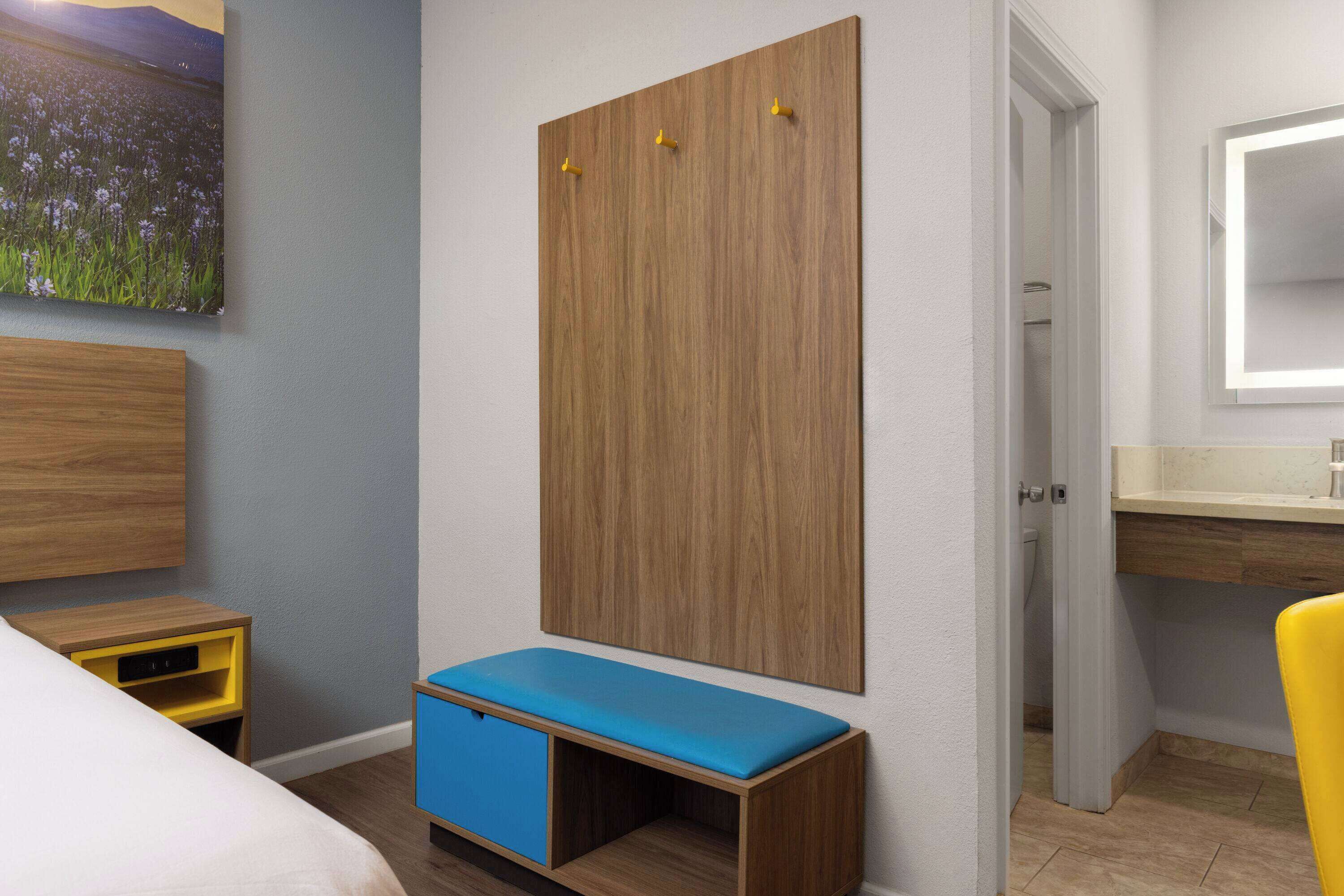A bedroom with a bed, a bedside table, a wooden wardrobe with hooks, a bench, a bathroom with a mirror, and a painting on the wall.