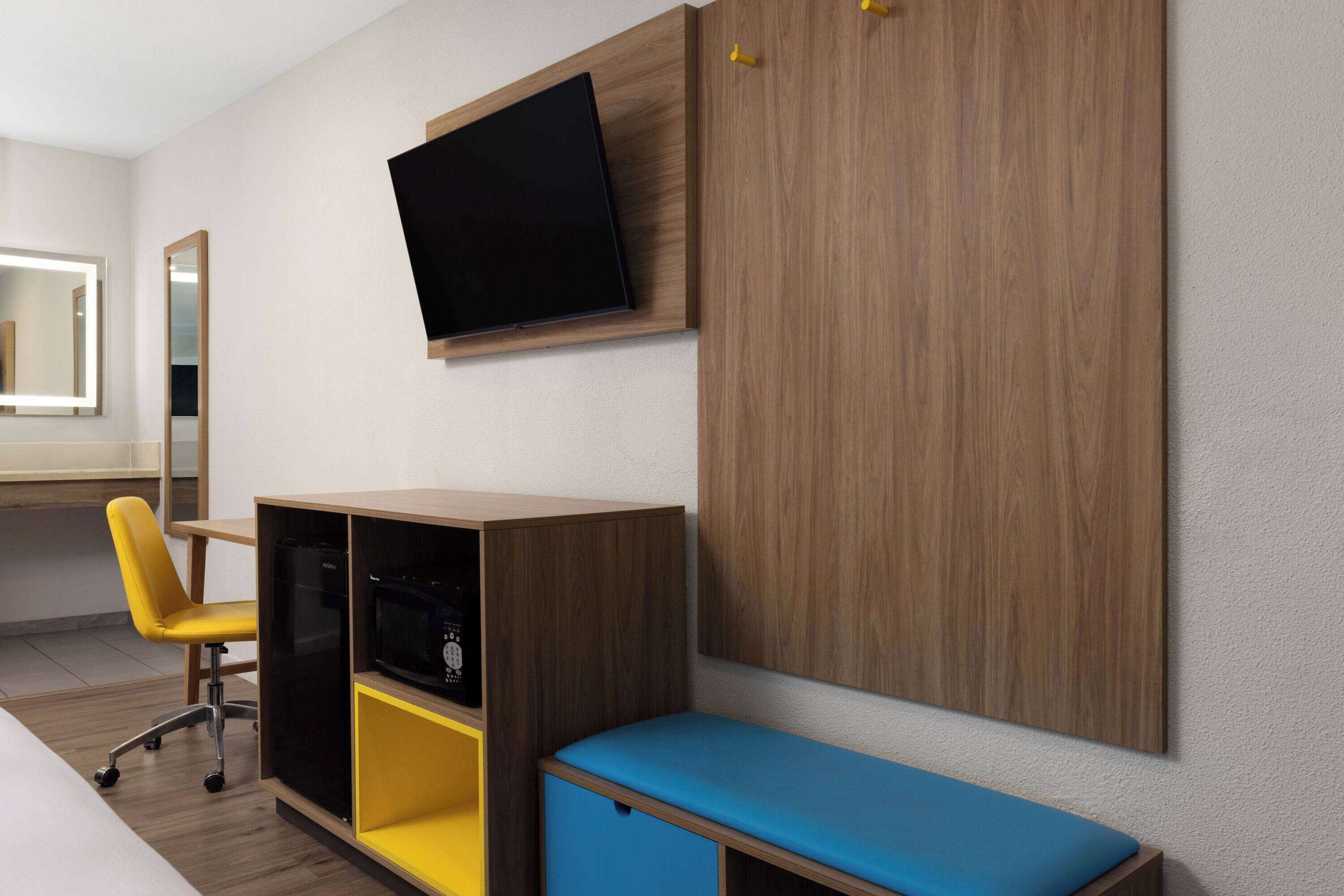 A modern hotel room with a wooden wardrobe, a flat-screen TV, a yellow chair, and a blue bench.