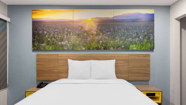 Habitación de hotel con un cuadro grande de un campo al atardecer, cabecera de madera, cama blanca y una mesita de noche con un teléfono.