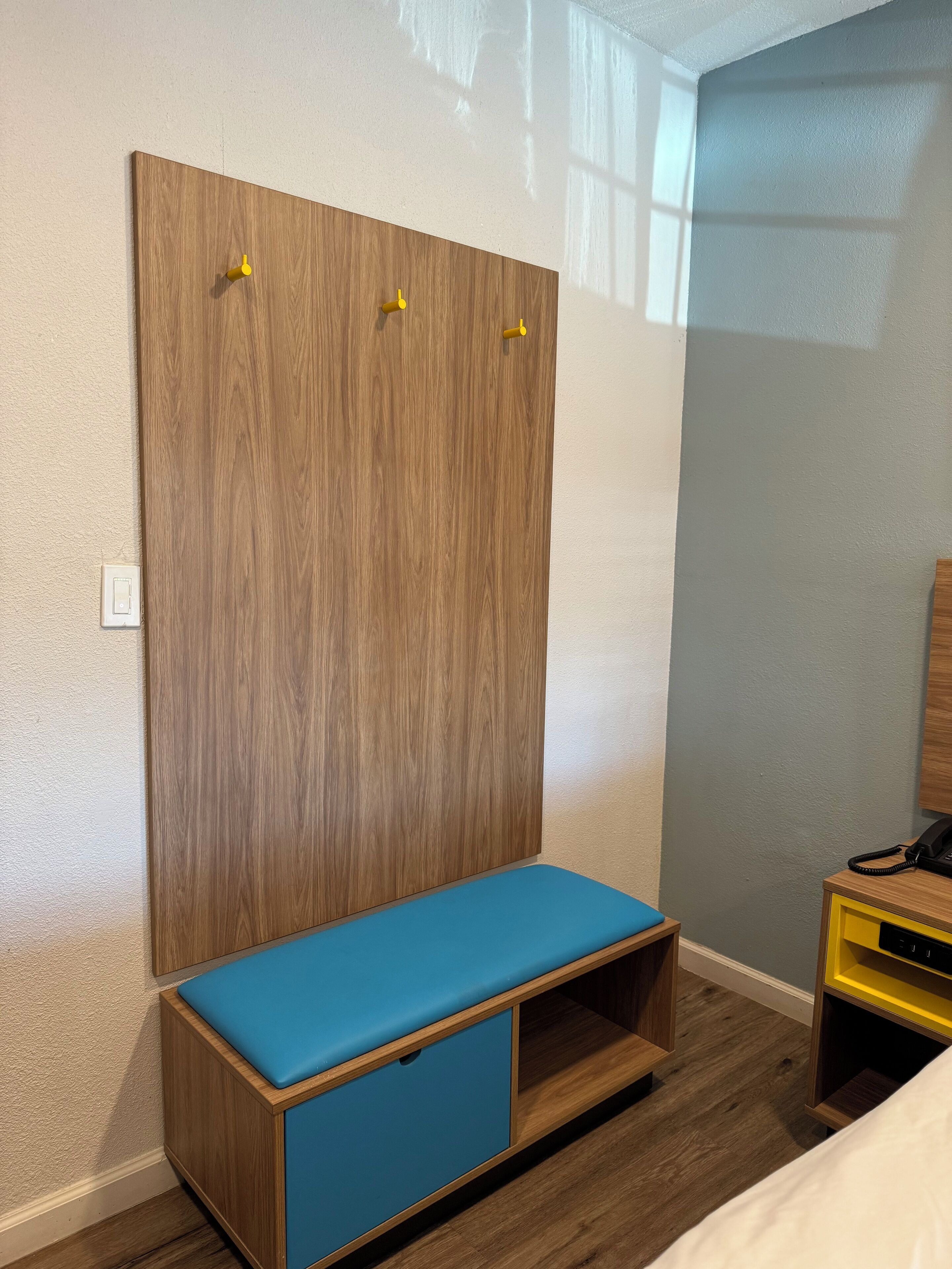 Un dormitorio con un armario de madera, un banco azul, una mesita de noche con un teléfono y un enchufe.