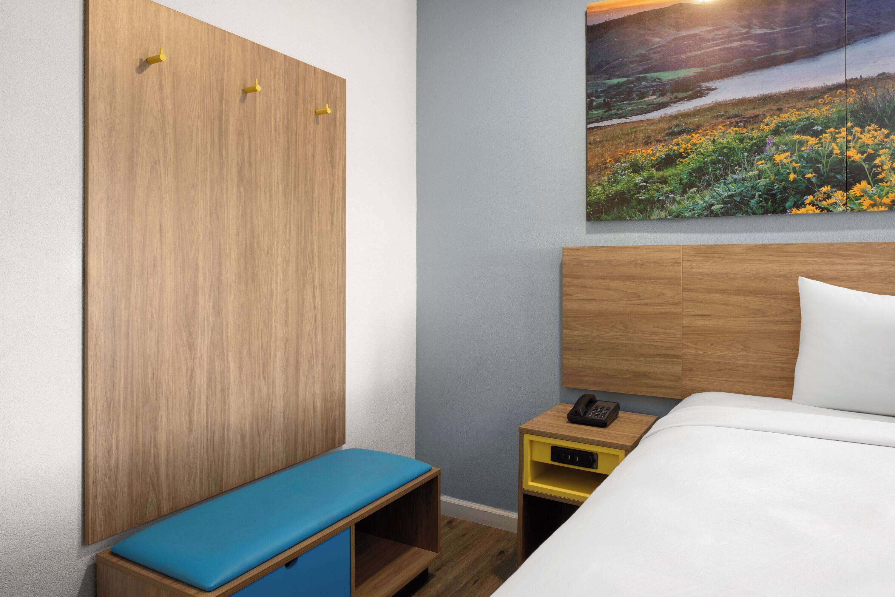 A hotel room with a bed, a wooden wardrobe, a bench, a bedside table with a phone, and a wall-mounted painting of a landscape.