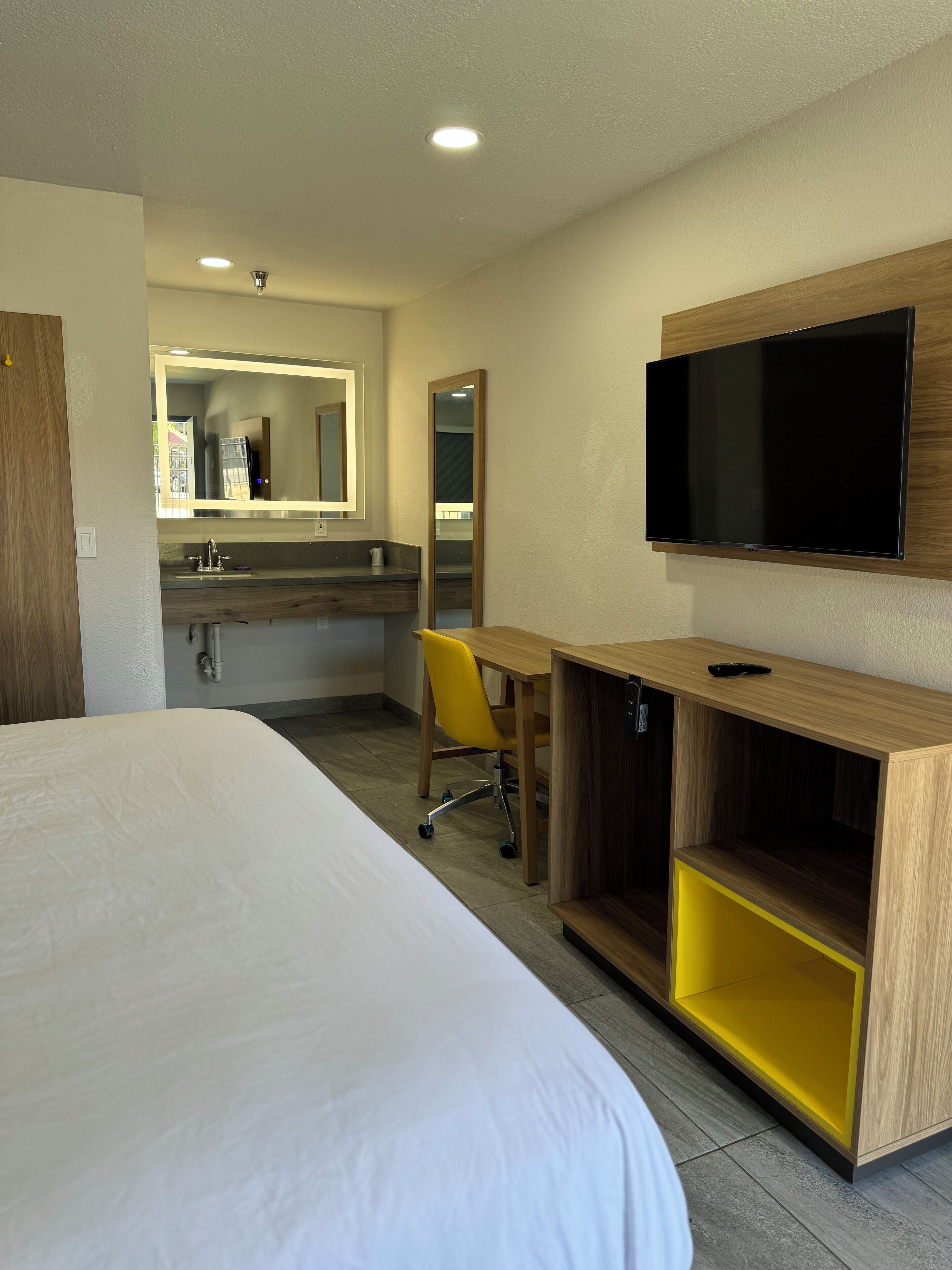 Habitación de hotel con una cama grande, un escritorio de madera con televisor, un espejo y un baño con lavamanos y espejo.