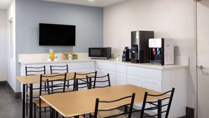 Cocina moderna con televisor de pantalla plana, microondas, cafetera y refrigerador. Seis mesas y sillas de madera.