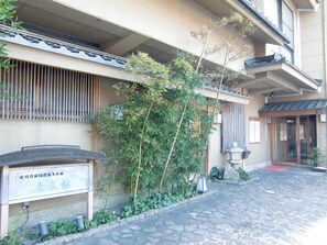 Un edificio japonés tradicional con entrada de adoquines, un letrero de madera y un pequeño jardín con plantas de bambú.