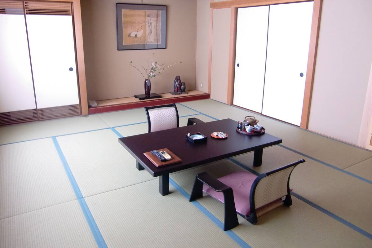 A traditional Japanese room with tatami flooring, a low table with cushions, a framed picture on the wall, and sliding doors.