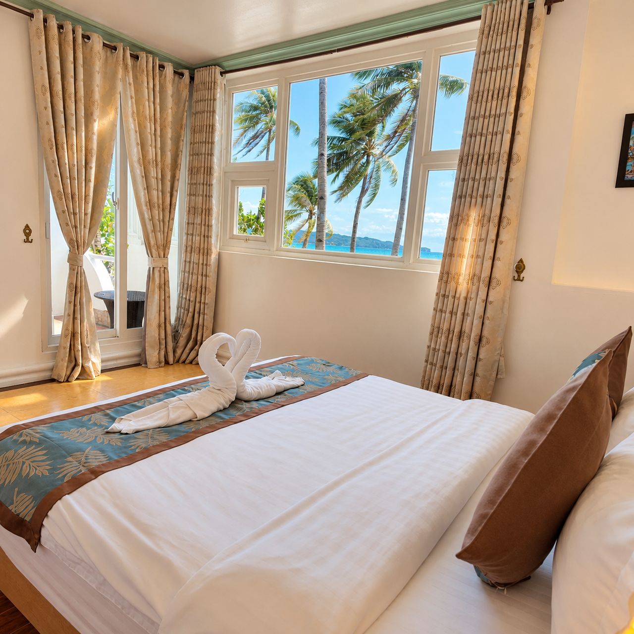 Una habitación de hotel con cama, cortinas, una ventana con vistas a palmeras y al mar, y una toalla sobre la cama.