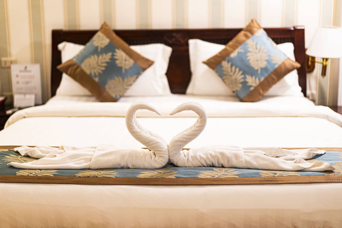 Quarto de hotel com cama bem arrumada, duas almofadas com padrão floral e toalha dobrada em forma de cisne sobre um tapete decorativo.