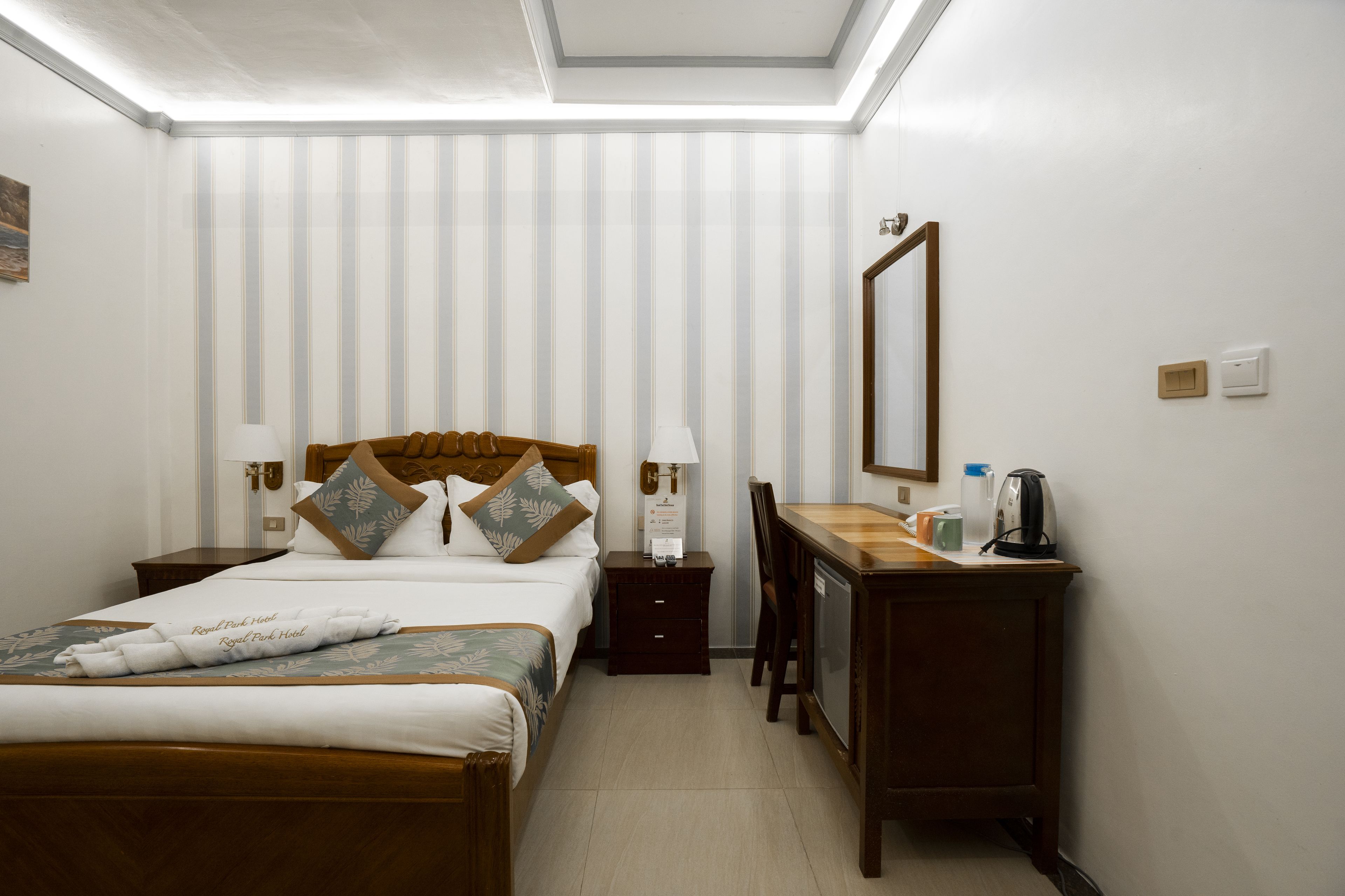 Une chambre d’hôtel comprenant un lit, un bureau, une chaise, une table de chevet, une lampe, un miroir et une fenêtre avec des stores.