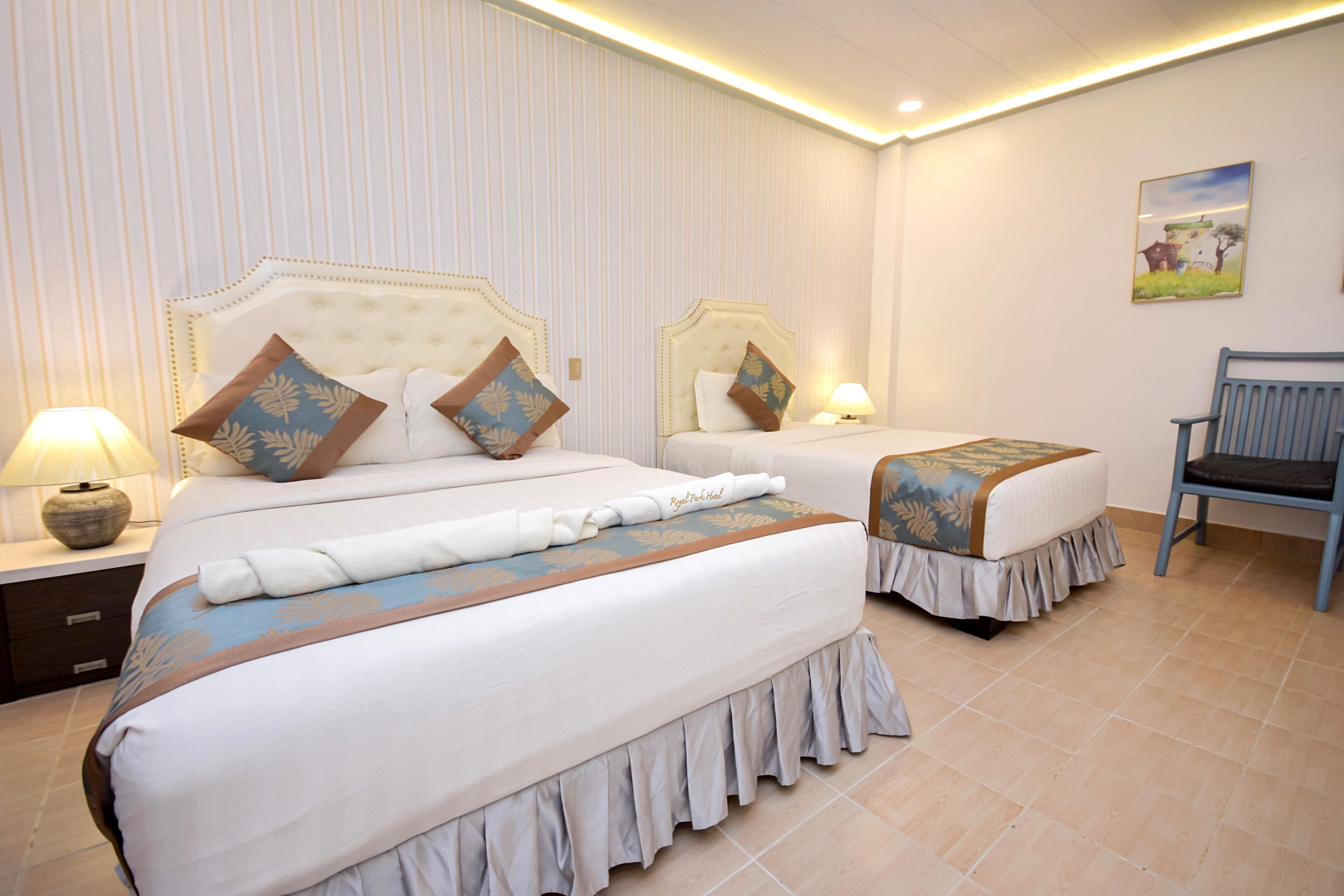 A hotel room with two beds, each with a headboard, bedside tables with lamps, and a bench.