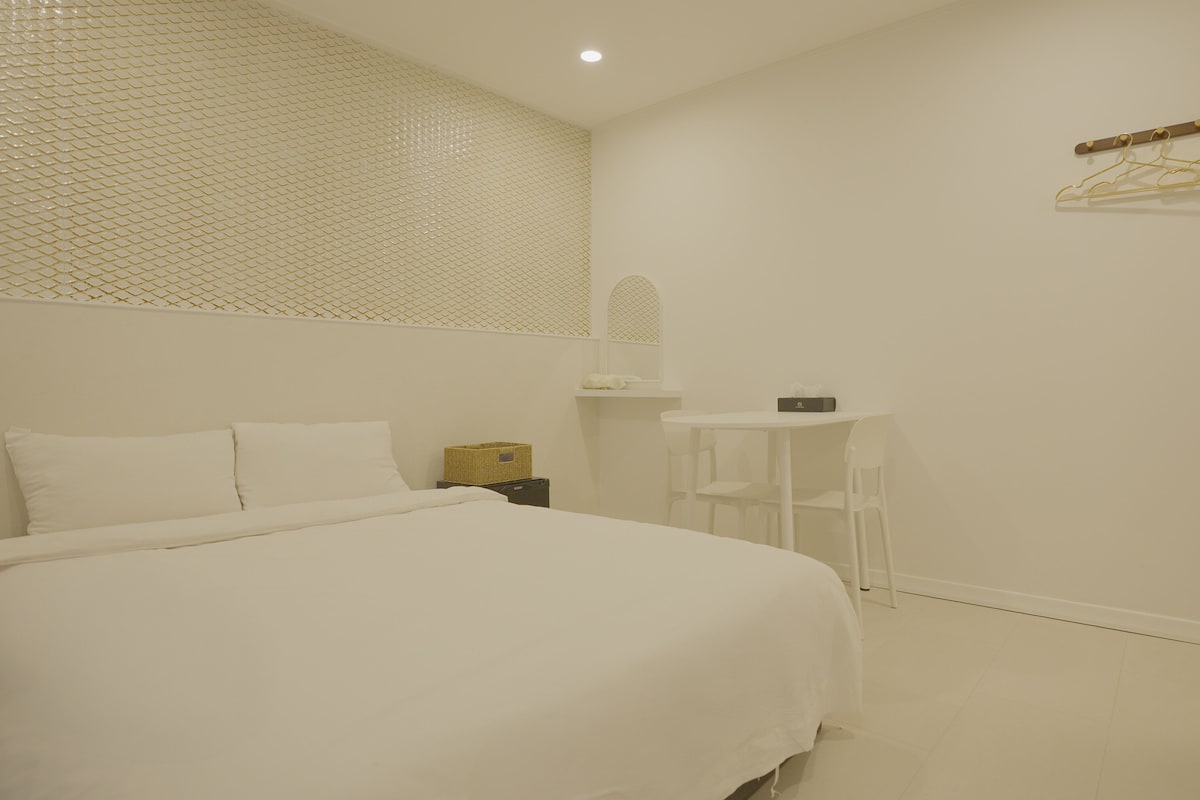A hotel room with a bed, a small table, a chair, a hanging clothes rack, and a wall-mounted shelf.