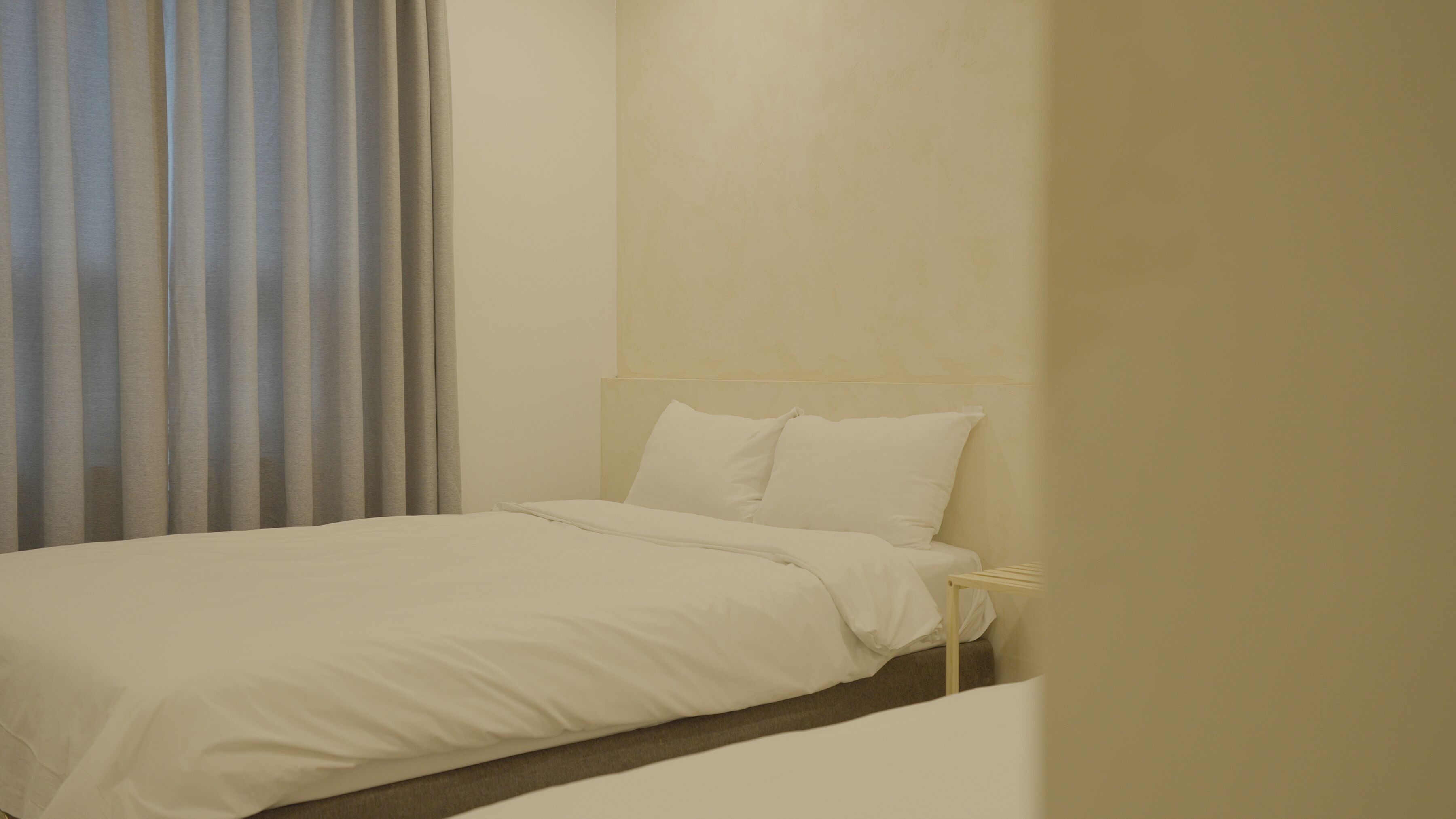 A neatly made bed with white bedding in a room with a plain wall and a window with vertical blinds.
