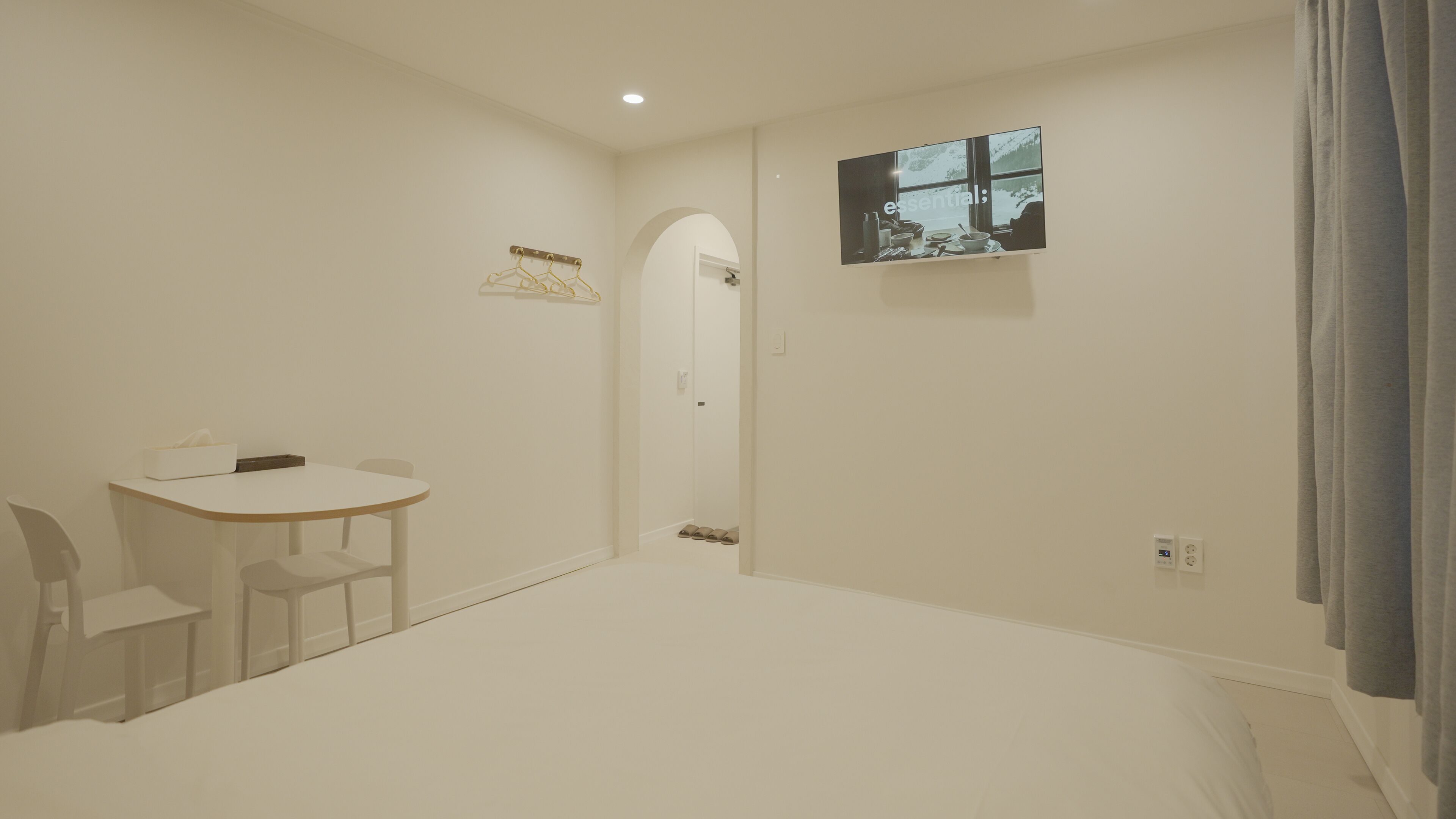 Een minimalistisch hotelkamer met een bed, een kleine tafel met stoelen, een aan de muur bevestigde televisie en een raam met gordijnen.