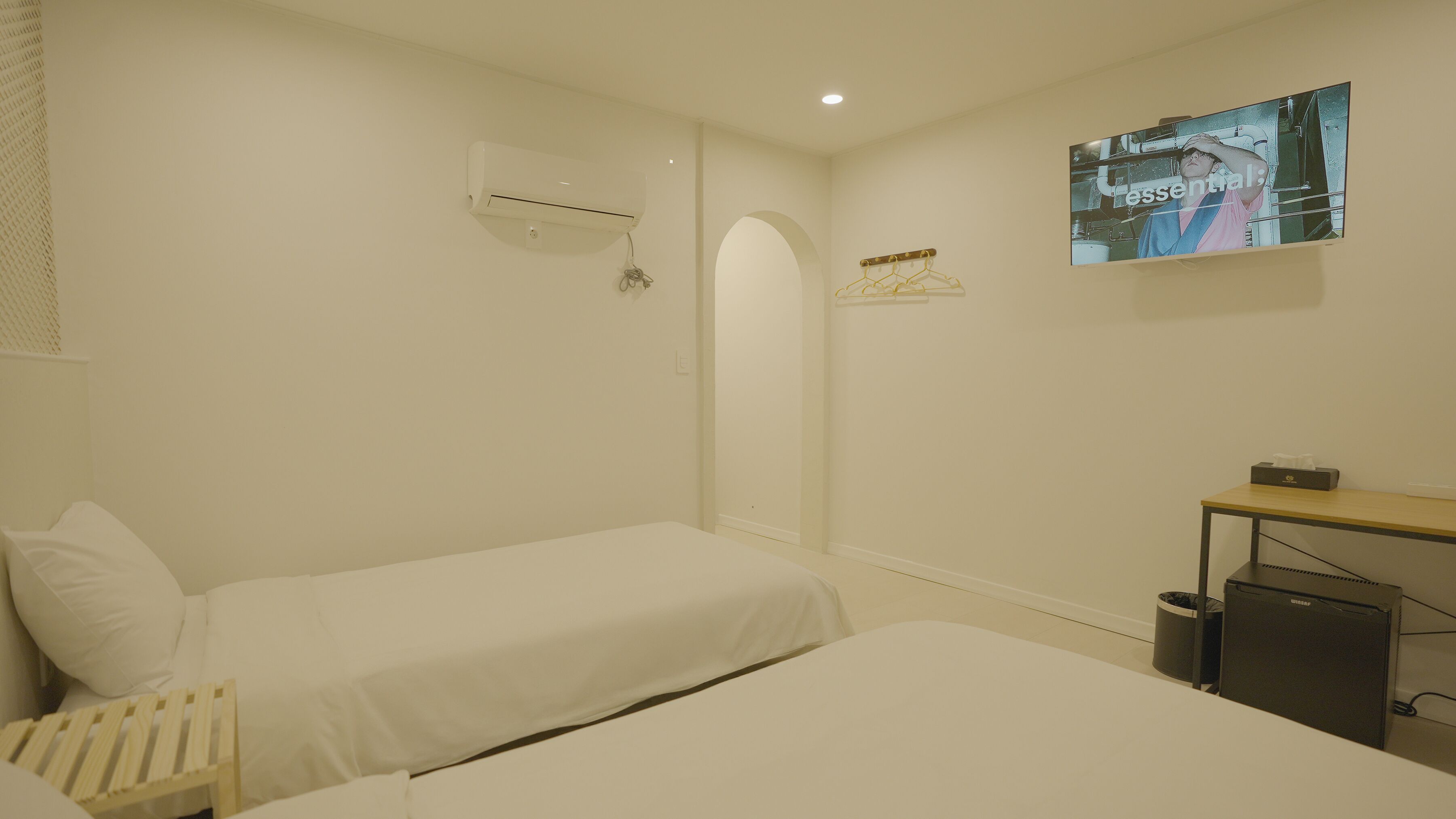 A hotel room with two beds, a desk, a chair, an air conditioning unit, a coat rack, and a wall-mounted television.