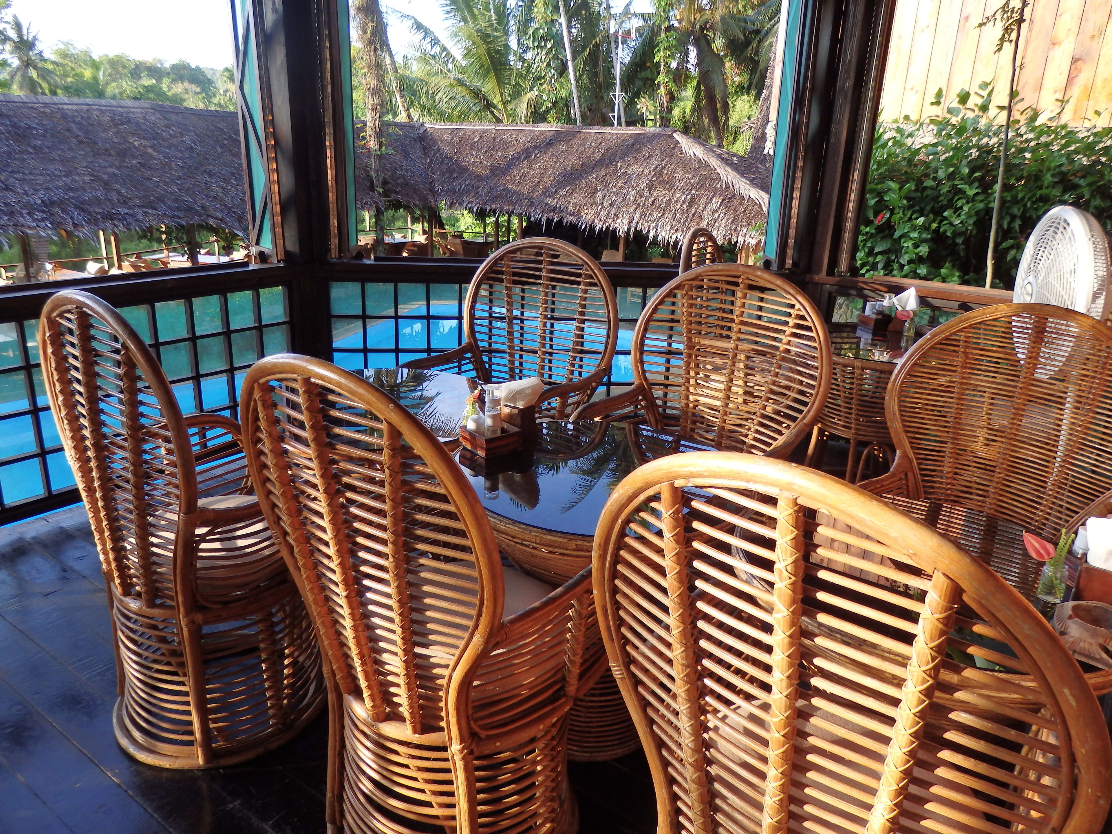 A tropical outdoor seating area with wicker chairs and a glass table, overlooking a pool and lush greenery.