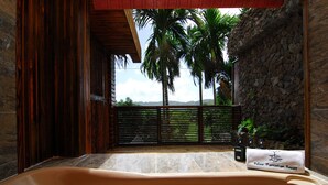 A spa room with a large bathtub, a stone wall, and a view of palm trees and mountains. - Palau Plantation Resort (Koror)