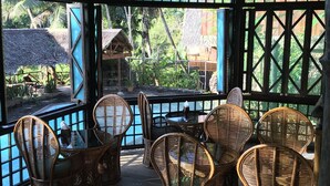 A covered outdoor seating area with wicker chairs and a table, overlooking a tropical garden and thatched-roof buildings. - Palau Plantation Resort (Koror)