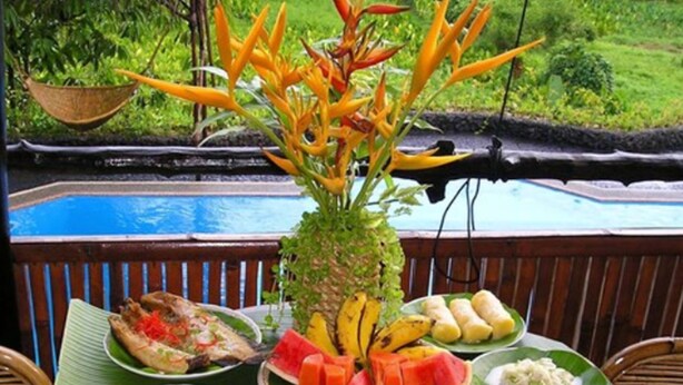 A table set with various dishes on banana leaves, a decorative plant, and a view of a pool. - Palau Plantation Resort (Koror)
