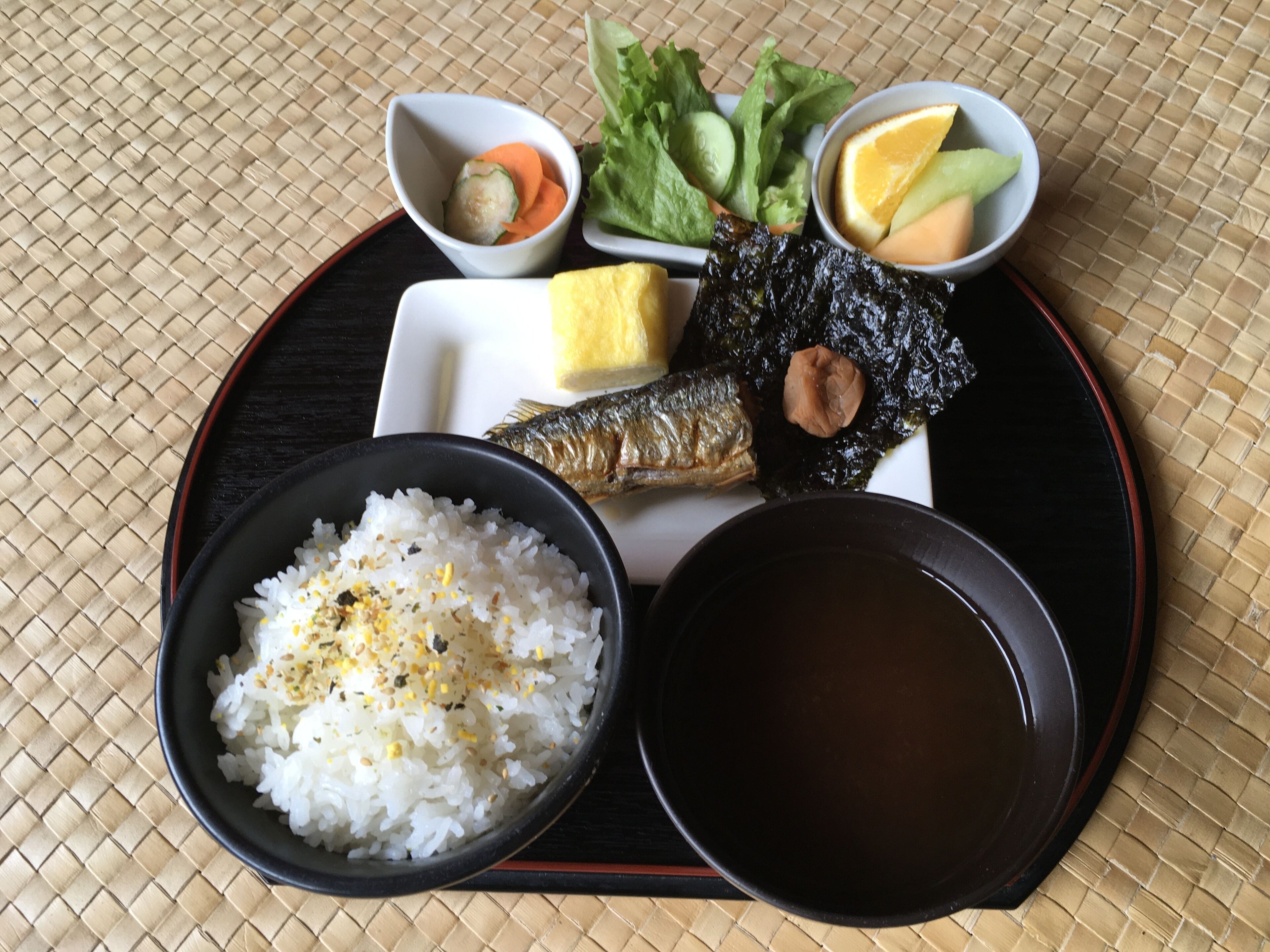 A meal consisting of rice, fish, vegetables, and a bowl of soup on a woven mat.