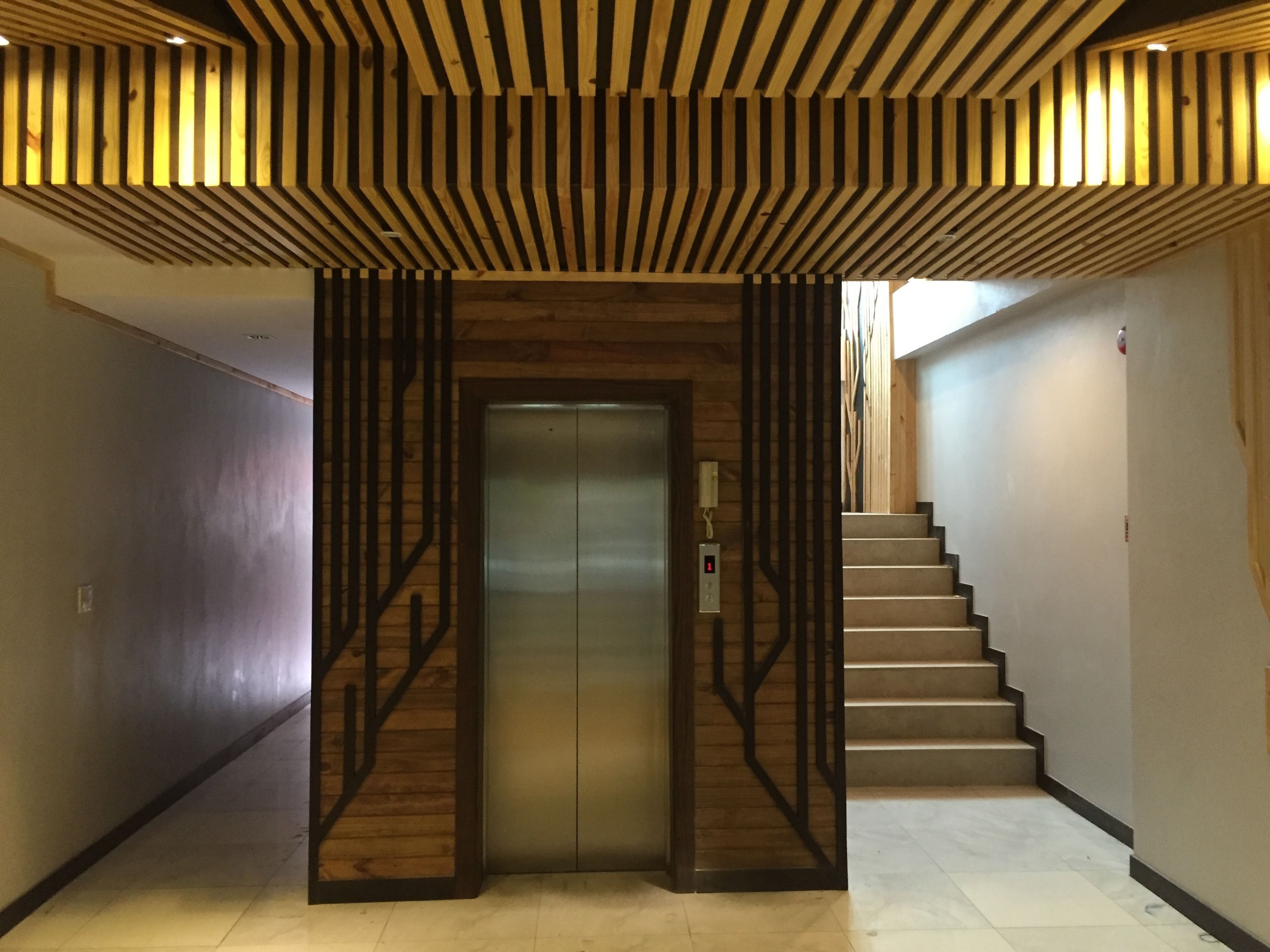 A modern elevator with a wooden panel design, a staircase with metal railings, and a plain wall on the left.