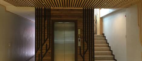 A modern elevator with a wooden panel design, a staircase with metal railings, and a plain wall on the left.