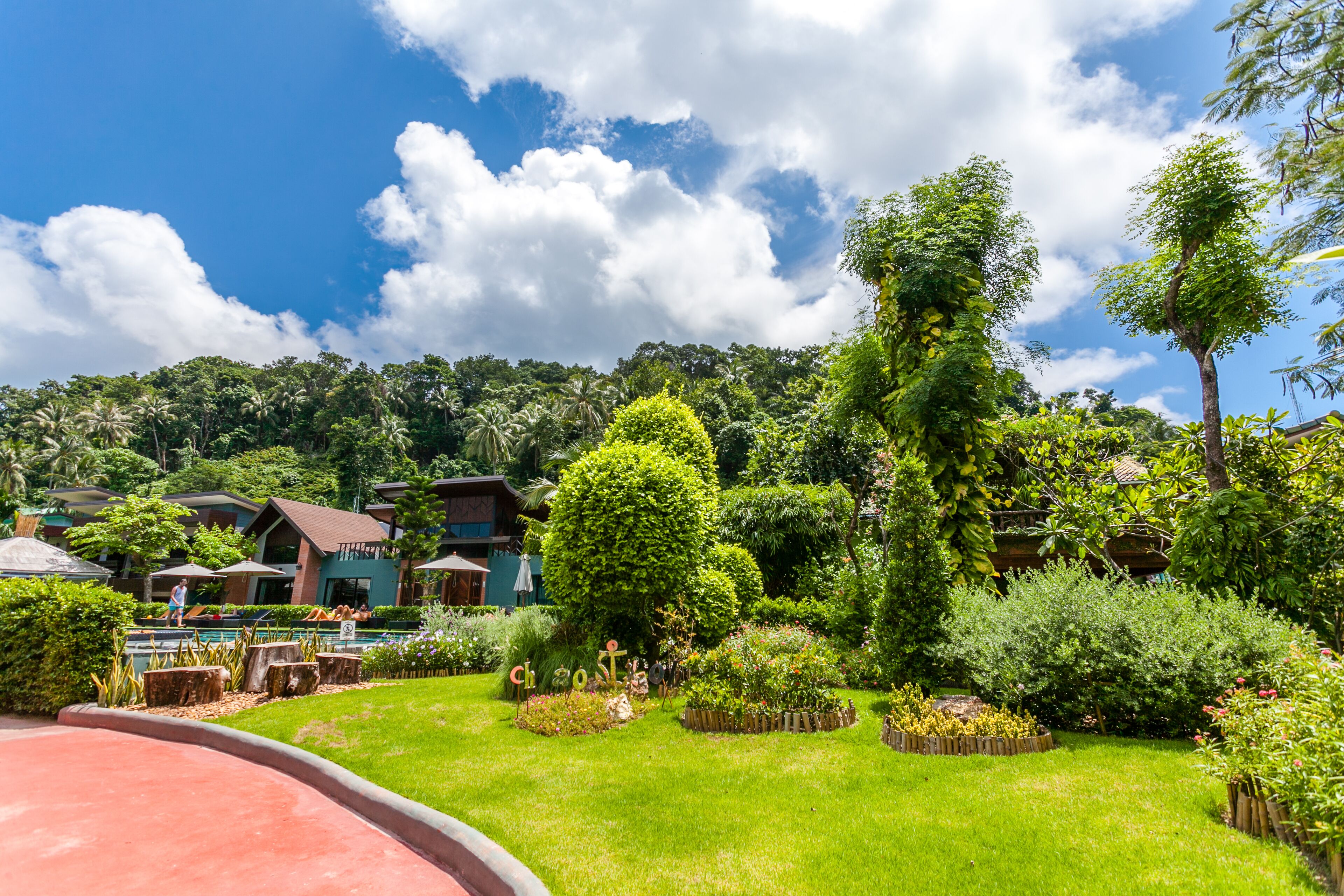 A lush garden with various plants and trees, a curved pathway, and buildings in the background.