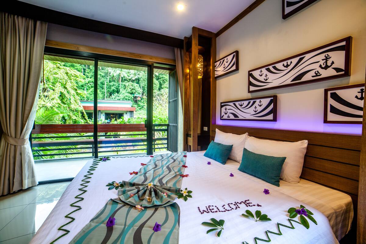 A hotel room with a large bed, a balcony with a view of greenery, and artwork on the walls.