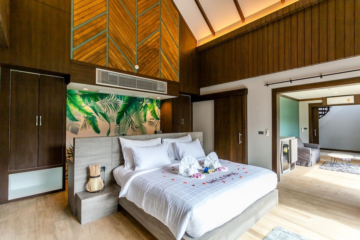 A modern bedroom with a large bed, wooden paneling, a tropical leaf mural, and a view of a dining area.